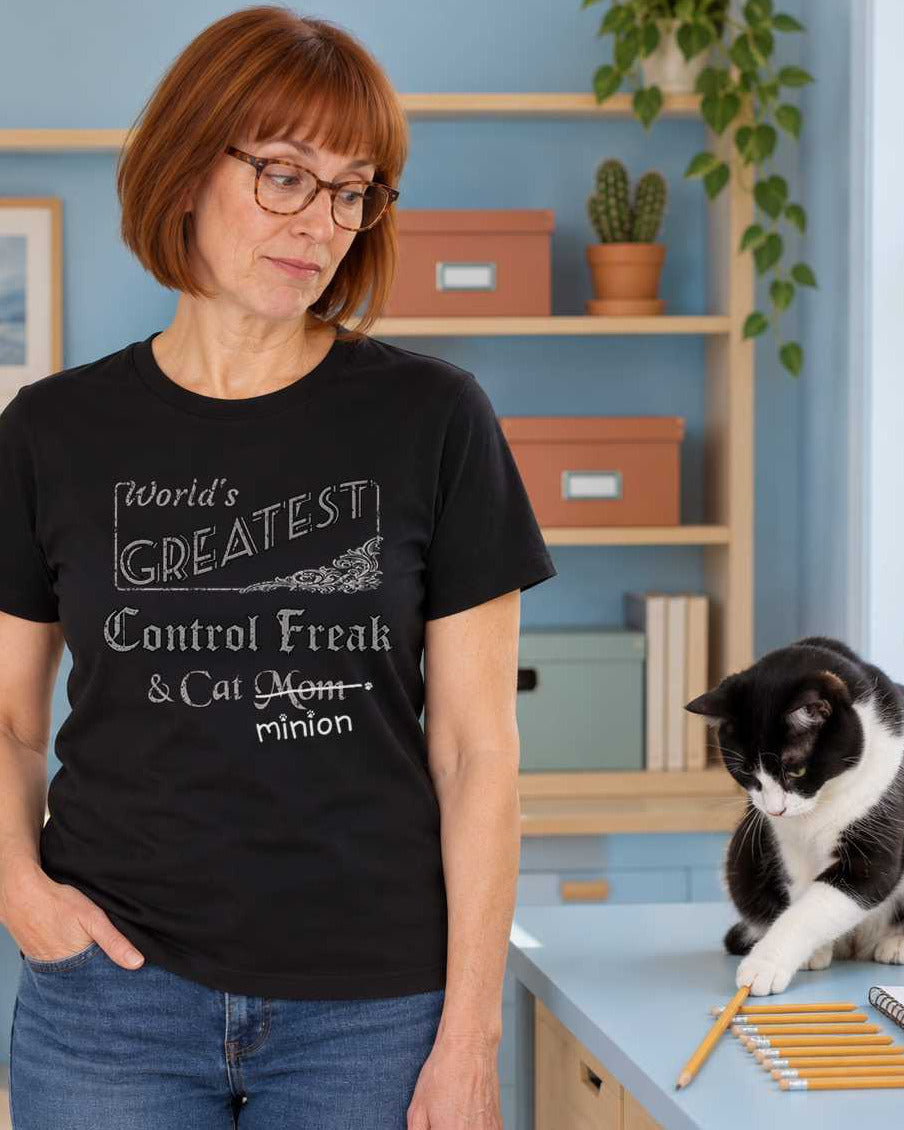 Woman watching a cat push a pencil out of an aligned row beside a desk, wearing a black crew-neck t-shirt that reads, “World’s Greatest Control Freak & Cat Mom” with “Mom” crossed out and “minion” handwritten below with paw prints.