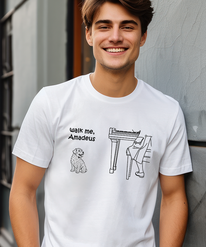 A smiling young man wearing a white graphic T-shirt with black text reading 'walk me, Amadeus' and line drawings of a dog and a harpsichord, standing against a gray textured wall.