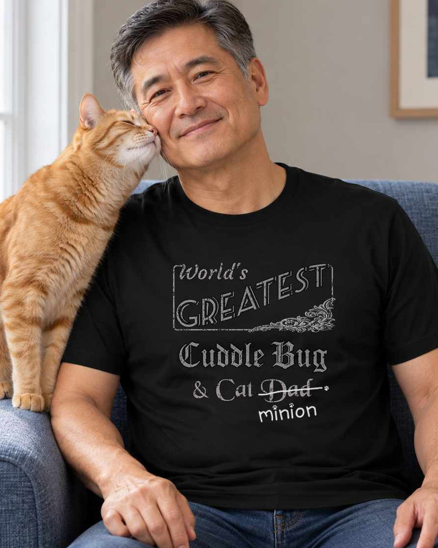 Man seated with a cat nuzzling their cheek from an armrest, wearing a black crew-neck t-shirt that reads, “World’s Greatest Cuddle Bug & Cat Dad” with “Dad” crossed out and “minion” handwritten below with paw prints.