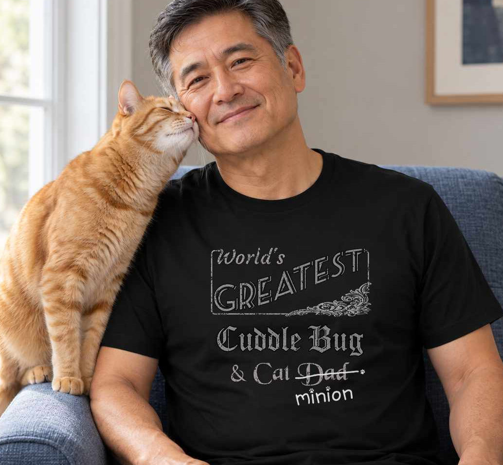 Man seated with a cat nuzzling their cheek from an armrest, wearing a black crew-neck t-shirt that reads, “World’s Greatest Cuddle Bug & Cat Dad” with “Dad” crossed out and “minion” handwritten below with paw prints.