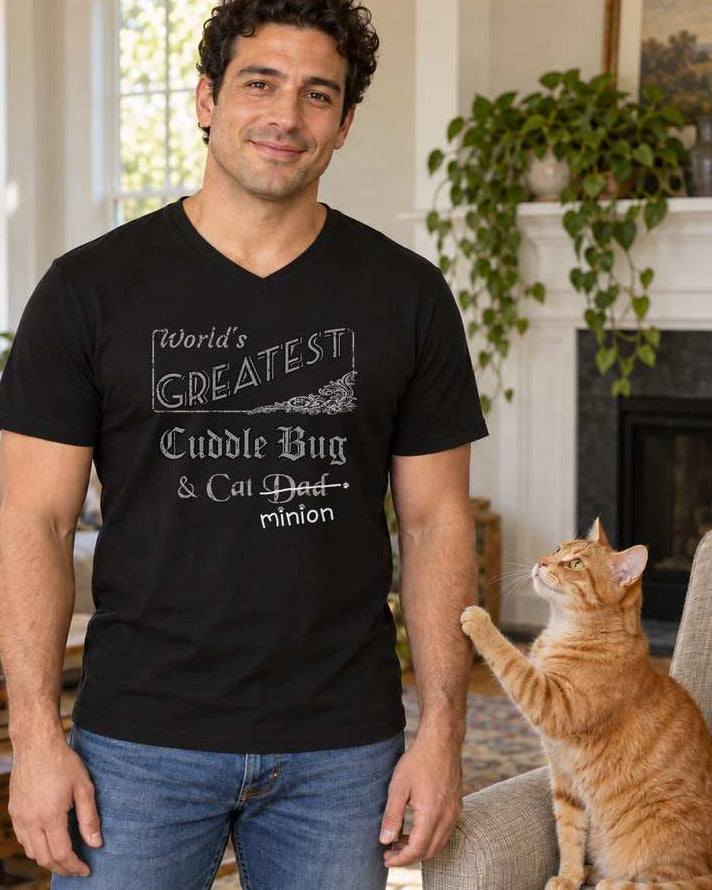 Man standing beside a ginger cat in a living room, wearing a black V-neck T-shirt reading “World’s Greatest Cuddle Bug & Cat Dad.” “Dad” is crossed out, with “minion” handwritten beneath it and surrounded by cat paw prints.