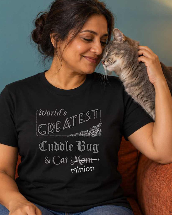 Woman seated with a cat nuzzling their cheek from an armrest, wearing a black crew-neck t-shirt that reads, “World’s Greatest Cuddle Bug & Cat Mom” with “Mom” crossed out and “minion” handwritten below with paw prints.