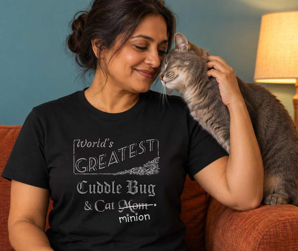 Woman seated with a cat nuzzling their cheek from an armrest, wearing a black crew-neck t-shirt that reads, “World’s Greatest Cuddle Bug & Cat Mom” with “Mom” crossed out and “minion” handwritten below with paw prints.