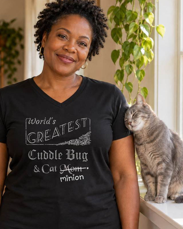 Woman beside a grey cat in a living room, wearing a black v-neck t-shirt that reads, “World’s Greatest Cuddle Bug & Cat Mom” with “Mom” crossed out and “minion” handwritten below with paw prints.