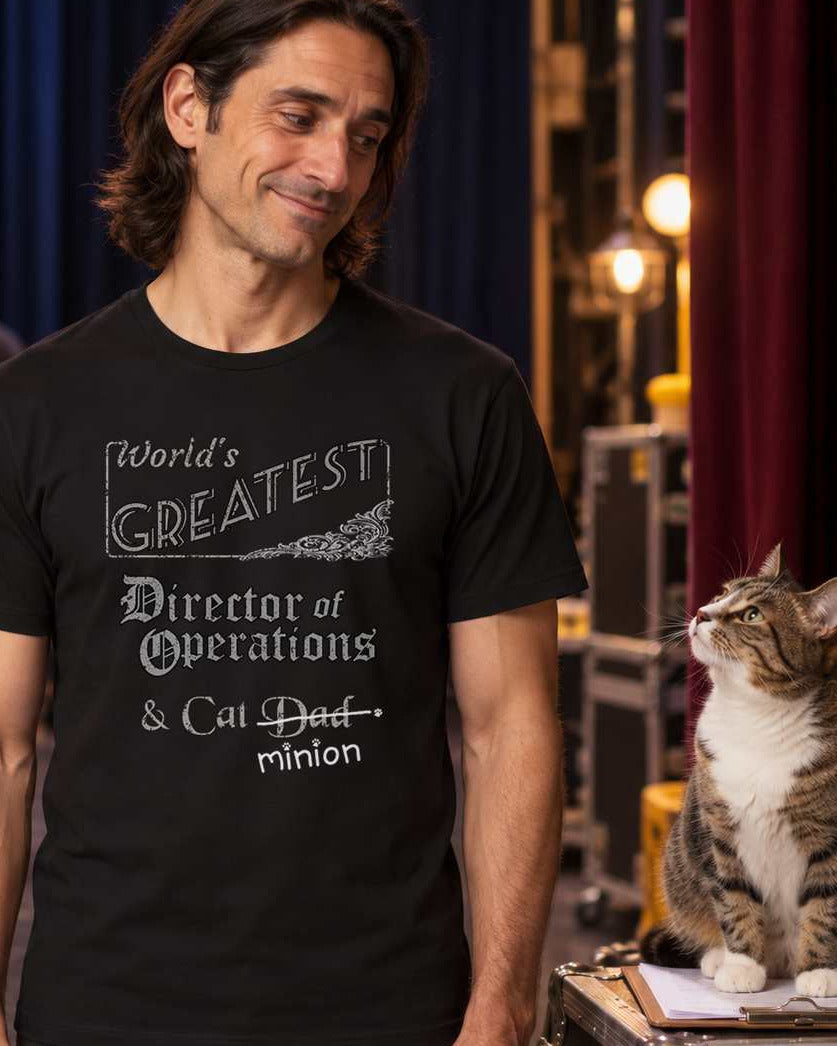 Man backstage with cat sitting on clipboard, wearing a black crew-neck t-shirt that reads, “World’s Greatest Director of Operations & Cat Dad” with “Dad” crossed out and “minion” handwritten below with paw prints.