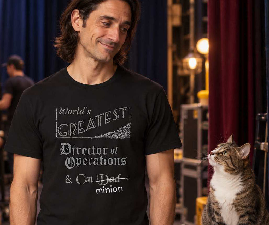 Man backstage with cat sitting on clipboard, wearing a black crew-neck t-shirt that reads, “World’s Greatest Director of Operations & Cat Dad” with “Dad” crossed out and “minion” handwritten below with paw prints.