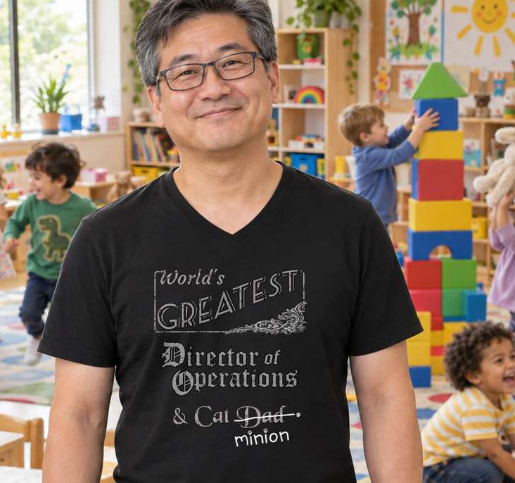 Man standing in a busy daycare classroom, wearing a black V-neck T-shirt reading “World’s Greatest Director of Operations & Cat Dad.” “Dad” is crossed out, with “minion” handwritten beneath it and surrounded by cat paw prints.