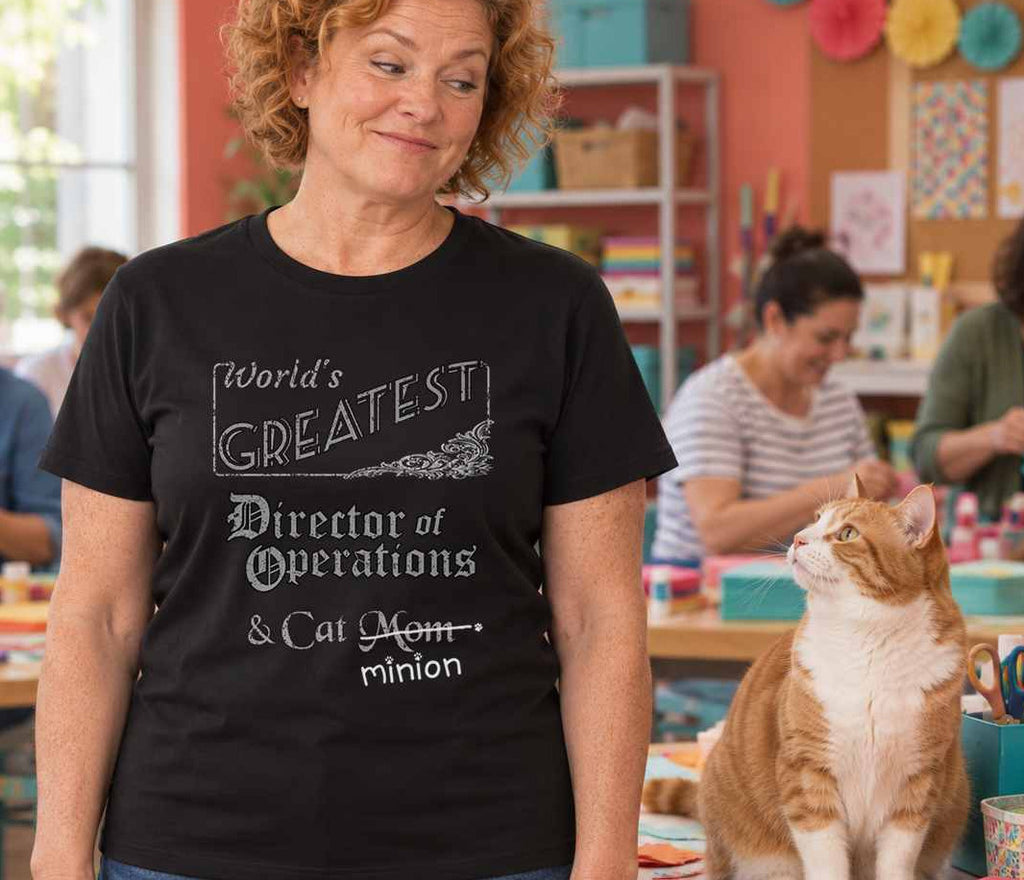 Woman in a craft room with cat on supplies, wearing a black crew-neck t-shirt that reads, “World’s Greatest Director of Operations & Cat Mom” with “Mom” crossed out and “minion” handwritten below with paw prints.