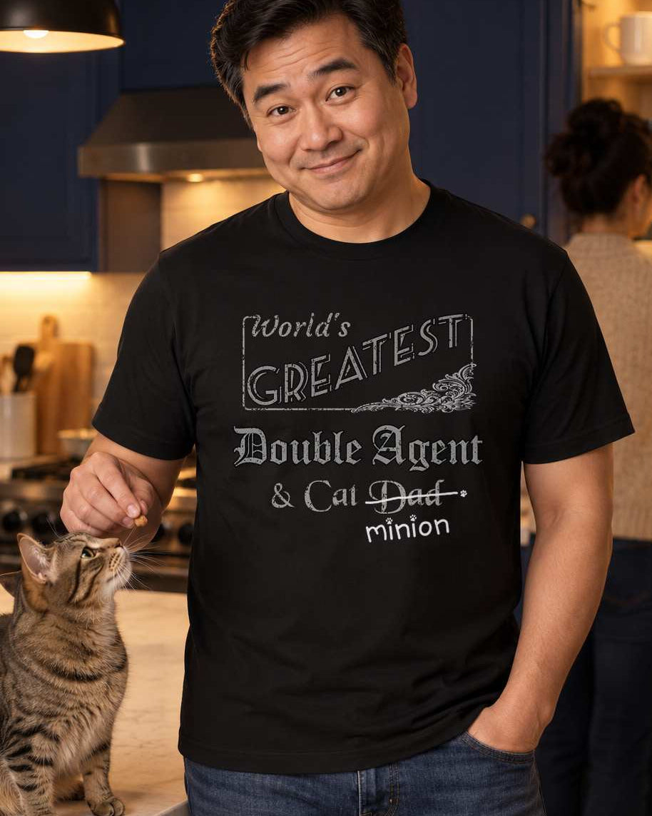 Man secretly offering a treat to a cat, wearing a black crew-neck t-shirt that reads, “World’s Greatest Double Agent & Cat Dad” with “Dad” crossed out and “minion” handwritten below with paw prints.