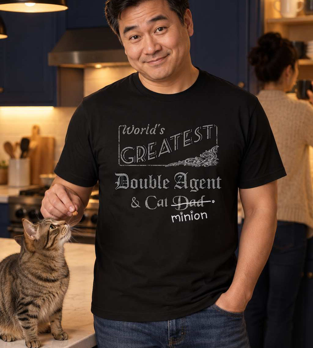 Man secretly offering a treat to a cat, wearing a black crew-neck t-shirt that reads, “World’s Greatest Double Agent & Cat Dad” with “Dad” crossed out and “minion” handwritten below with paw prints.