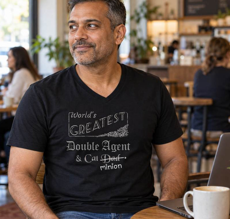 Man sitting at a café table with people behind him, wearing a black V-neck T-shirt reading “World’s Greatest Double Agent & Cat Dad.” “Dad” is crossed out, with “minion” handwritten beneath it and surrounded by cat paw prints.