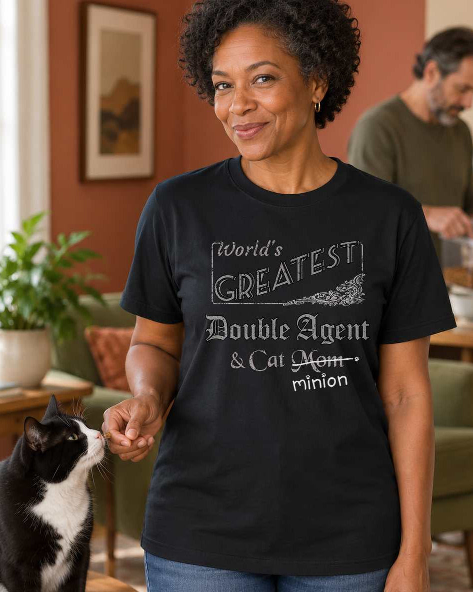 Woman secretly offering a treat to a cat, wearing a black crew-neck t-shirt that reads, “World’s Greatest Double Agent & Cat Mom” with “Mom” crossed out and “minion” handwritten below with paw prints.
