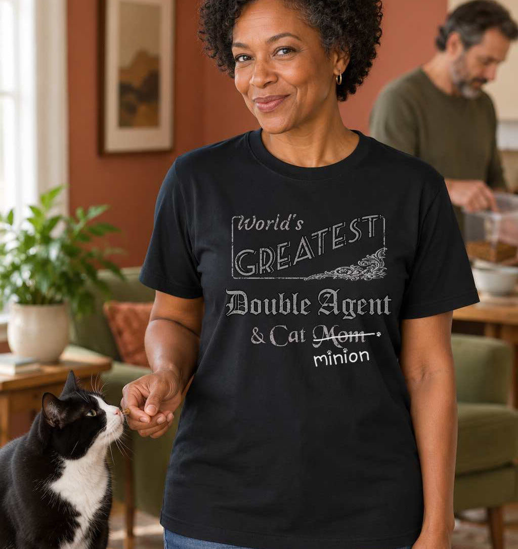 Woman secretly offering a treat to a cat, wearing a black crew-neck t-shirt that reads, “World’s Greatest Double Agent & Cat Mom” with “Mom” crossed out and “minion” handwritten below with paw prints.