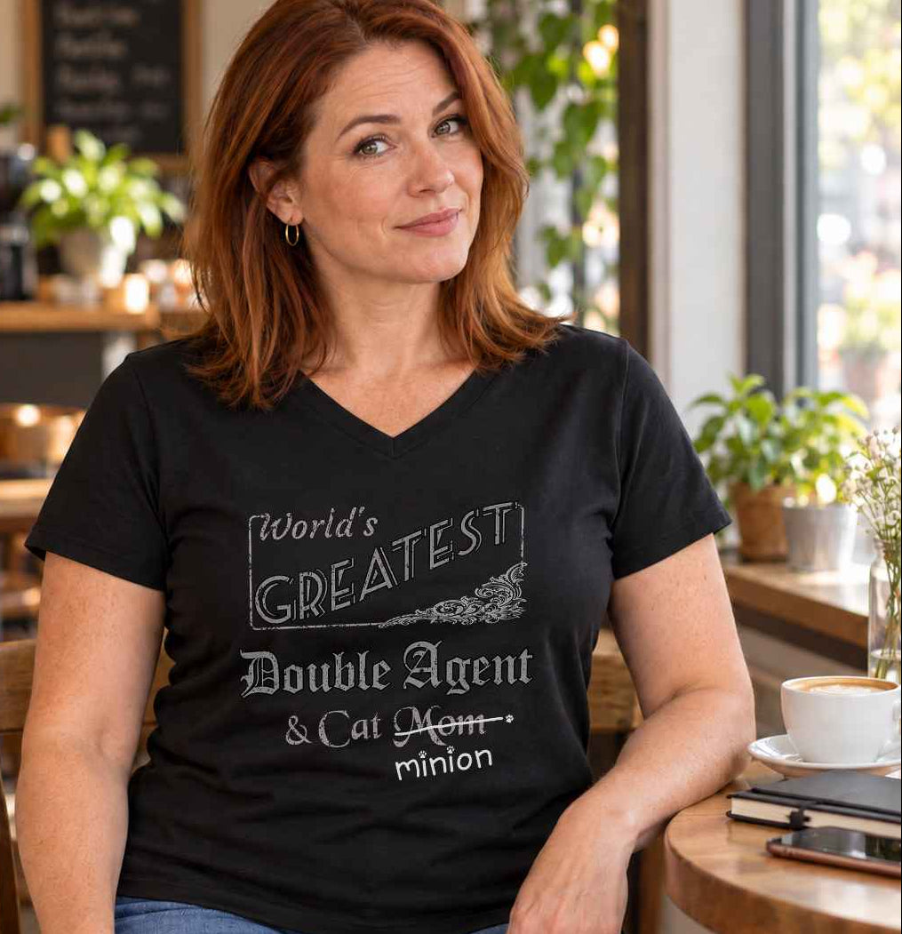 Woman sitting at a café table with people behind her, wearing a black V-neck T-shirt reading “World’s Greatest Double Agent & Cat Mom.” “Mom” is crossed out, with “minion” handwritten beneath it and surrounded by cat paw prints.