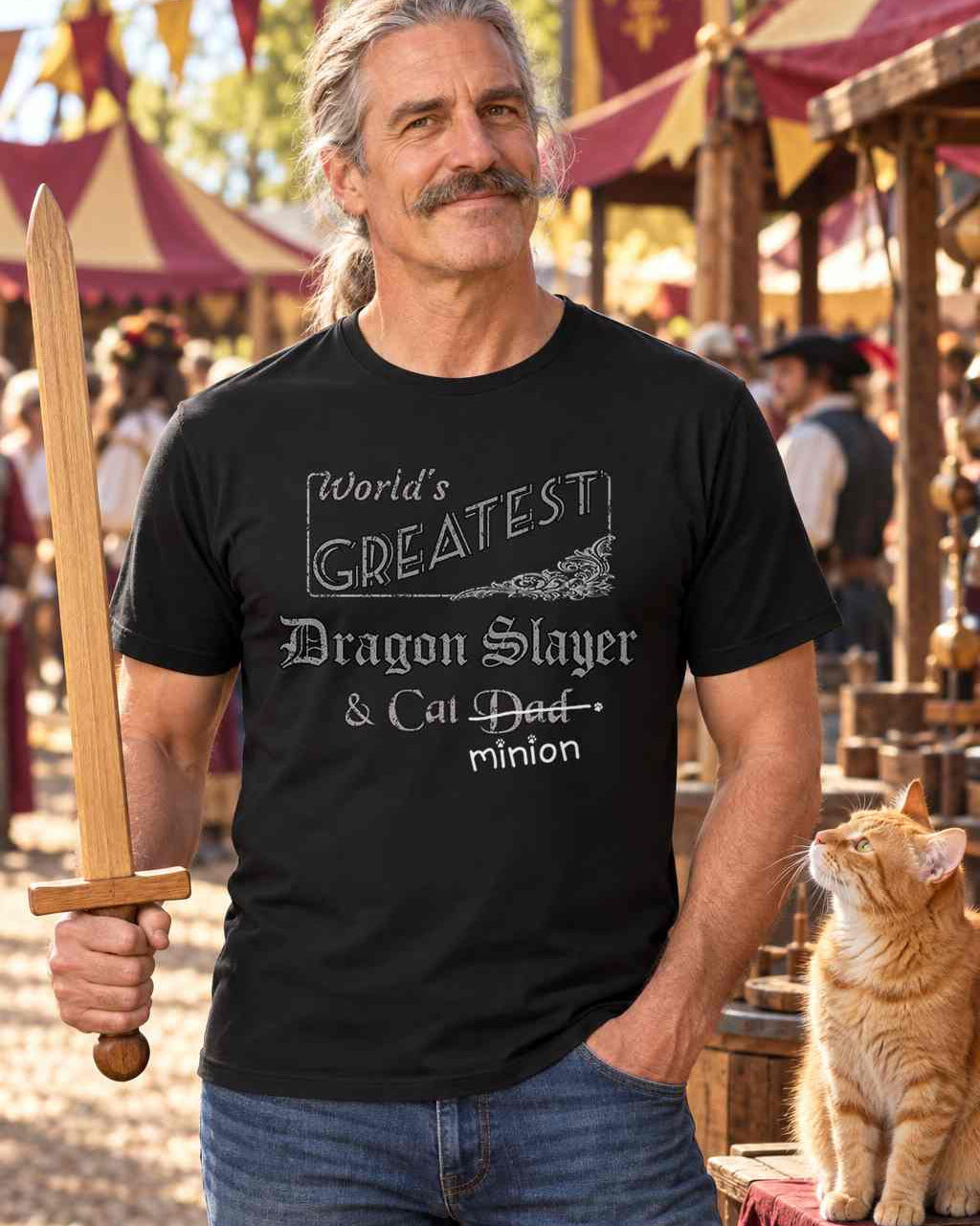 Man at a Renaissance faire holding a wooden practice sword beside a ginger cat, wearing a black crew-neck t-shirt that reads, “World’s Greatest Dragon Slayer & Cat Dad” with “Dad” crossed out and “minion” handwritten below with paw prints.