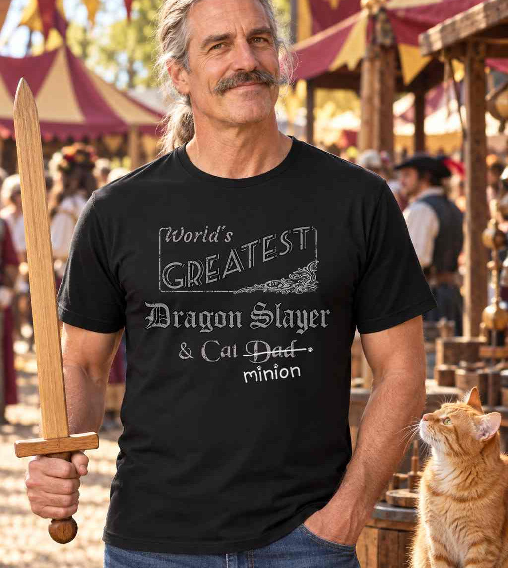 Man at a Renaissance faire holding a wooden practice sword beside a ginger cat, wearing a black crew-neck t-shirt that reads, “World’s Greatest Dragon Slayer & Cat Dad” with “Dad” crossed out and “minion” handwritten below with paw prints.