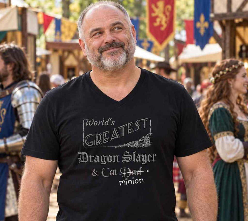 Man standing among children at a party, wearing a black V-neck T-shirt reading “World’s Greatest Dragon Slayer & Cat Dad.” “Dad” is crossed out, with “minion” handwritten beneath it and surrounded by cat paw prints.