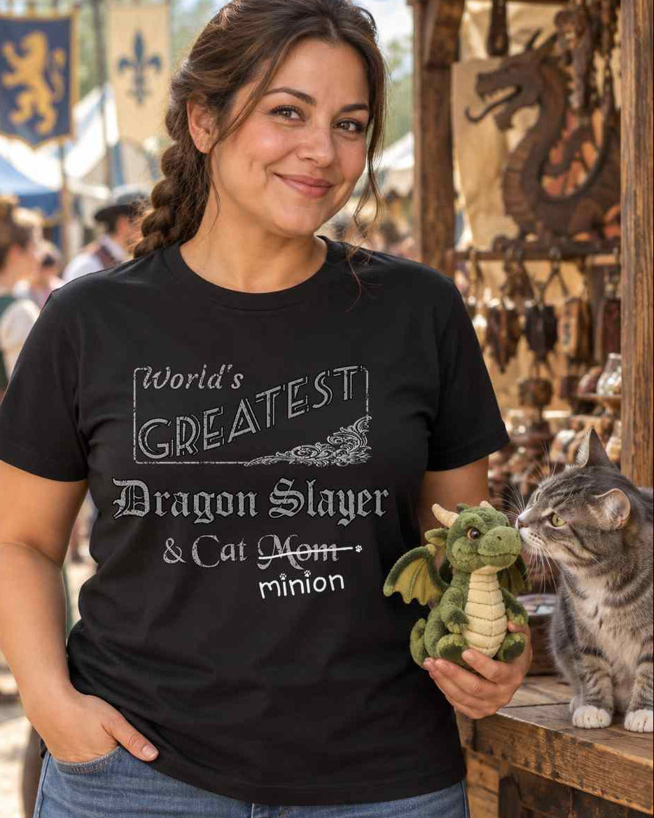 Woman at a Renaissance faire holding a toy dragon beside a tabby cat, wearing a black crew-neck t-shirt that reads, “World’s Greatest Dragon Slayer & Cat Mom” with “Mom” crossed out and “minion” handwritten below with paw prints.