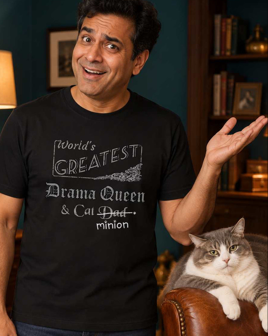 Man gesturing toward a cat occupying an armchair, wearing a black crew-neck t-shirt that reads, “World’s Greatest Drama Queen & Cat Dad” with “Dad” crossed out and “minion” handwritten below with paw prints.
