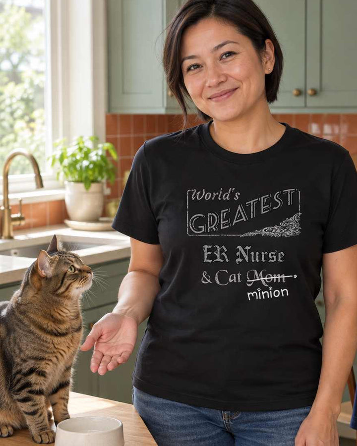 Woman standing beside a waiting cat and food bowl in a kitchen, wearing a black crew-neck t-shirt that reads, “World’s Greatest ER Nurse & Cat Mom” with “Mom” crossed out and “minion” handwritten below with paw prints.