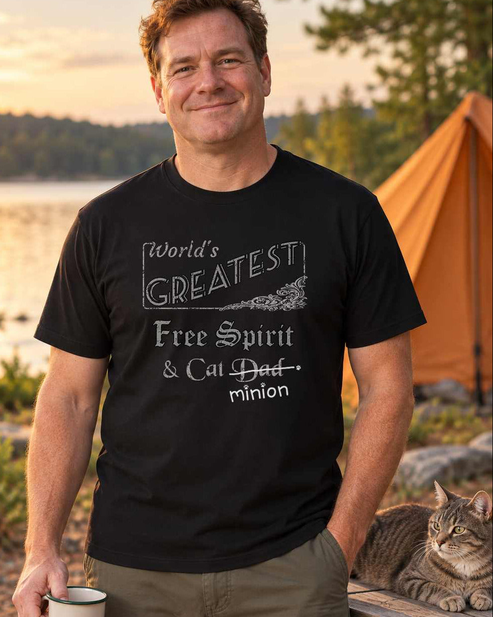 Man at a lakeside campsite holding an enamel mug beside a cat on a picnic table, wearing a black crew-neck t-shirt that reads, “World’s Greatest Free Spirit & Cat Dad” with “Dad” crossed out and “minion” handwritten below with paw prints.