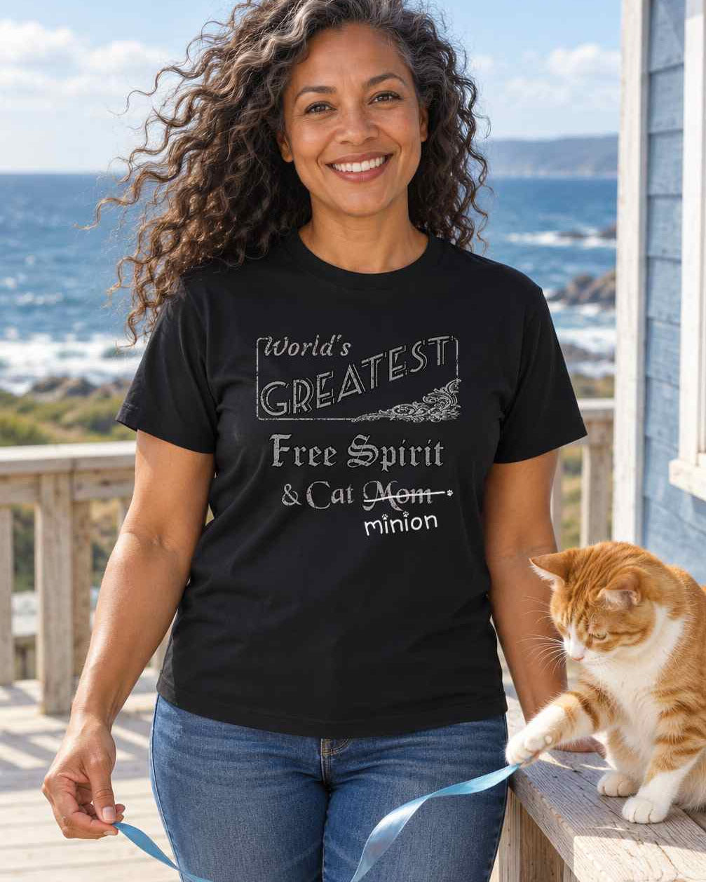 Woman on a breezy coastal deck holding a ribbon for a cat, wearing a black crew-neck t-shirt that reads, “World’s Greatest Free Spirit & Cat Mom” with “Mom” crossed out and “minion” handwritten below with paw prints.