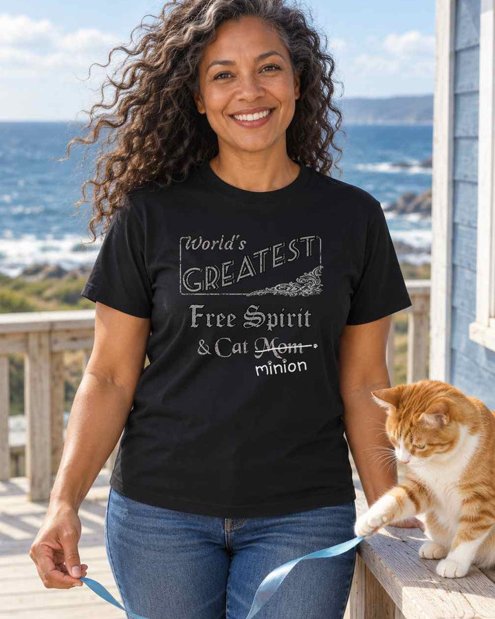 Woman on a breezy coastal deck holding a ribbon for a cat, wearing a black crew-neck t-shirt that reads, “World’s Greatest Free Spirit & Cat Mom” with “Mom” crossed out and “minion” handwritten below with paw prints.