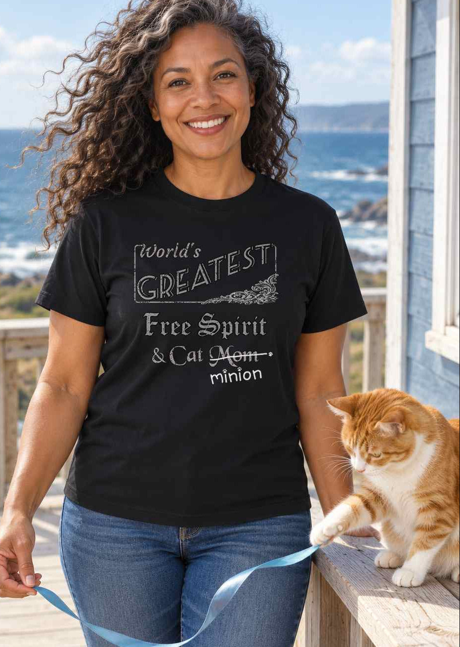 Woman on a breezy coastal deck holding a ribbon for a cat, wearing a black crew-neck t-shirt that reads, “World’s Greatest Free Spirit & Cat Mom” with “Mom” crossed out and “minion” handwritten below with paw prints.