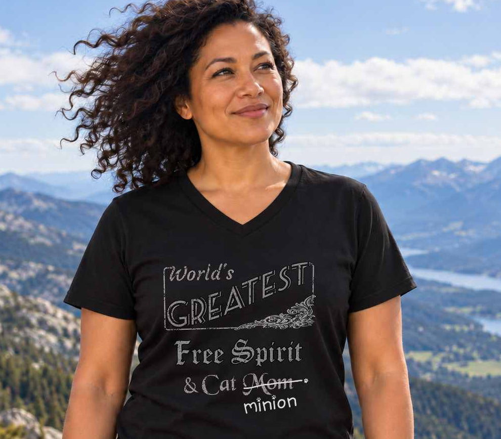 Woman standing in mountainous countryside, wearing a black V-neck T-shirt reading “World’s Greatest Free Spirit & Cat Mom.” “Mom” is crossed out, with “minion” handwritten beneath it and surrounded by cat paw prints.