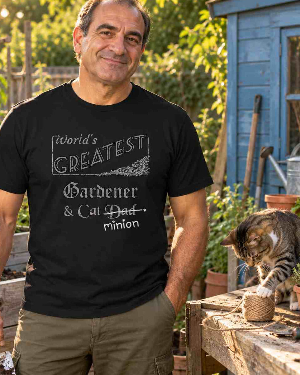 Man holding a trowel in a vegetable garden beside a cat investigating twine, wearing a black crew-neck t-shirt that reads, “World’s Greatest Gardener & Cat Dad” with “Dad” crossed out and “minion” handwritten below with paw prints.