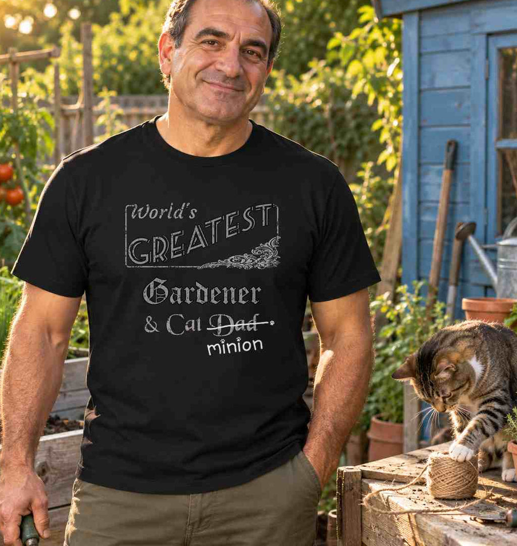 Man holding a trowel in a vegetable garden beside a cat investigating twine, wearing a black crew-neck t-shirt that reads, “World’s Greatest Gardener & Cat Dad” with “Dad” crossed out and “minion” handwritten below with paw prints.