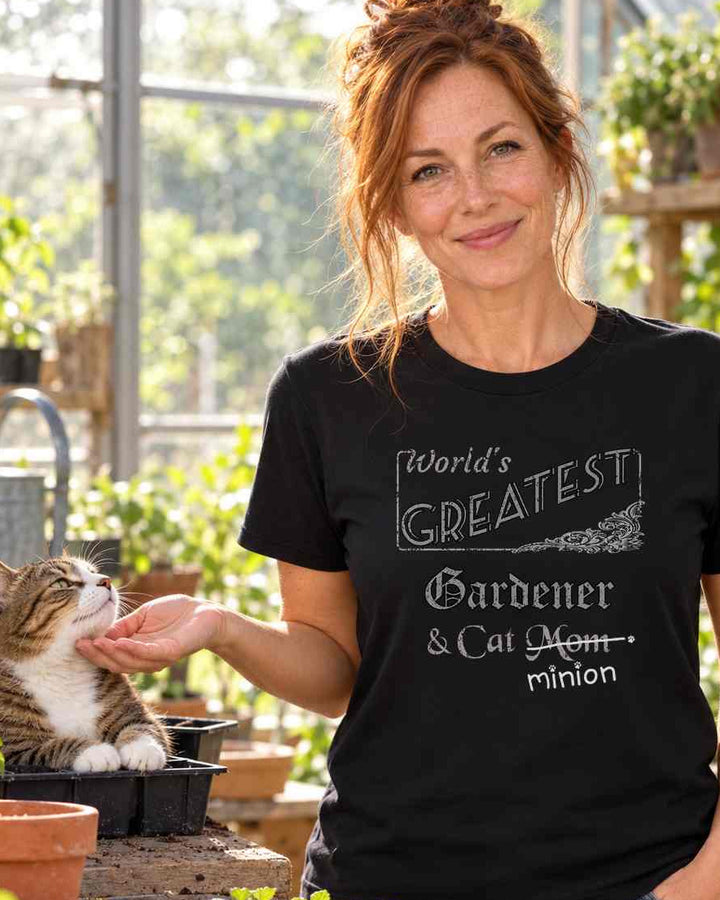Woman petting a cat in a seed tray at a greenhouse potting bench, wearing a black crew-neck t-shirt that reads, “World’s Greatest Gardener & Cat Mom” with “Mom” crossed out and “minion” handwritten below with paw prints.
