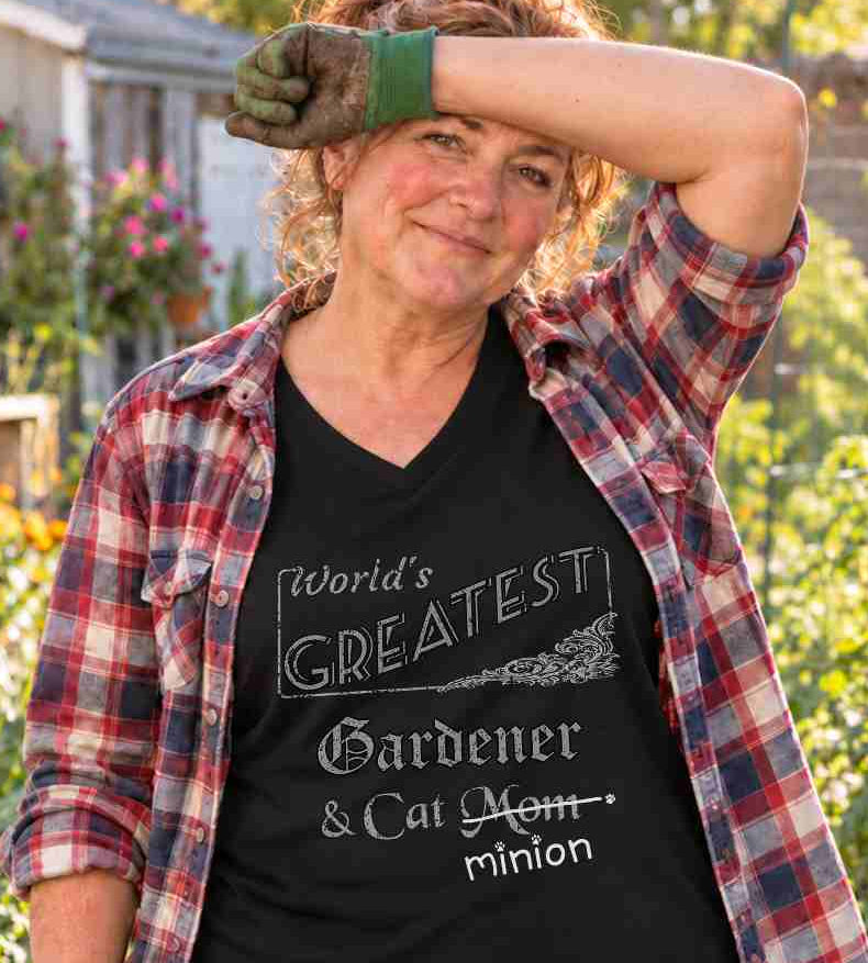 Woman pausing while gardening, with a cat nearby, wearing a black V-neck T-shirt reading “World’s Greatest Gardener & Cat Mom.” “Mom” is crossed out, with “minion” handwritten beneath it and surrounded by cat paw prints.