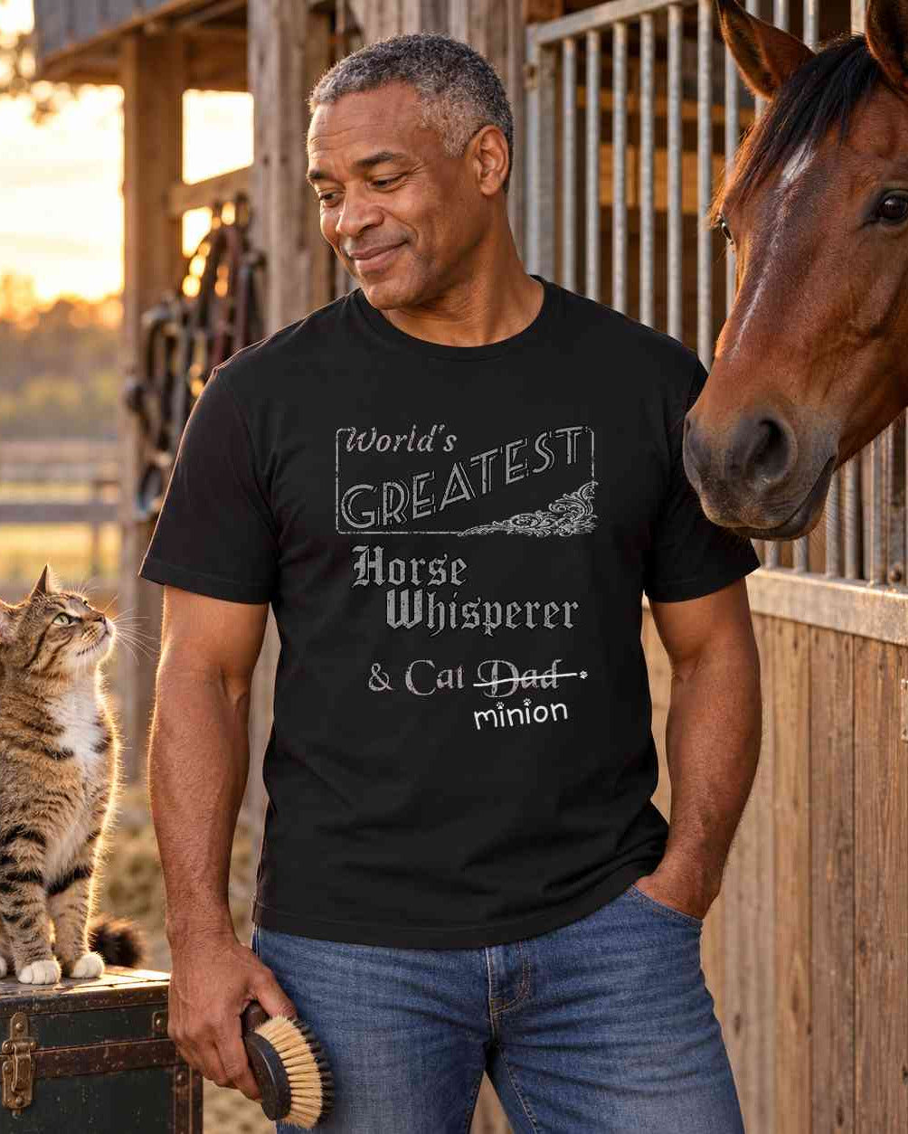 Man holding a grooming brush beside a horse at a stable and a cat on a tack trunk, wearing a black crew-neck t-shirt that reads, “World’s Greatest Horse Whisperer & Cat Dad” with “Dad” crossed out and “minion” handwritten below with paw prints.