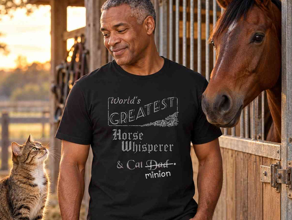 Man holding a grooming brush beside a horse at a stable and a cat on a tack trunk, wearing a black crew-neck t-shirt that reads, “World’s Greatest Horse Whisperer & Cat Dad” with “Dad” crossed out and “minion” handwritten below with paw prints.