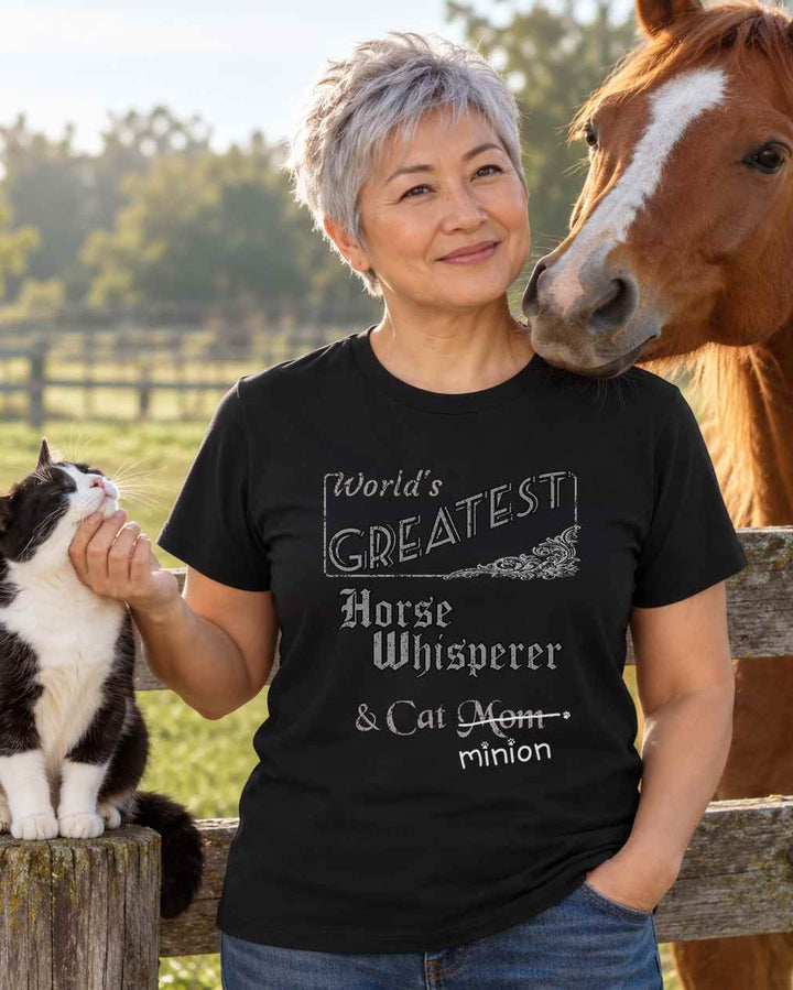 Woman scratching a cat’s chin beside a horse at a paddock fence, wearing a black crew-neck t-shirt that reads, “World’s Greatest Horse Whisperer & Cat Mom” with “Mom” crossed out and “minion” handwritten below with paw prints.
