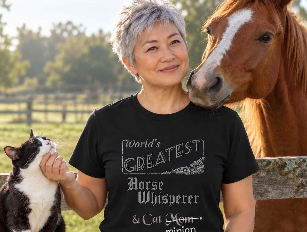 Woman scratching a cat’s chin beside a horse at a paddock fence, wearing a black crew-neck t-shirt that reads, “World’s Greatest Horse Whisperer & Cat Mom” with “Mom” crossed out and “minion” handwritten below with paw prints.