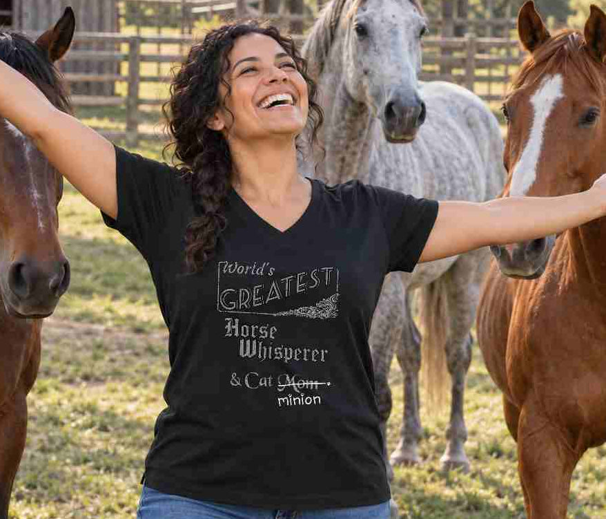 Woman standing with her arms outstretched among horses, wearing a black V-neck T-shirt reading “World’s Greatest Horse Whisperer & Cat Mom.” “Mom” is crossed out, with “minion” handwritten beneath it and surrounded by cat paw prints.