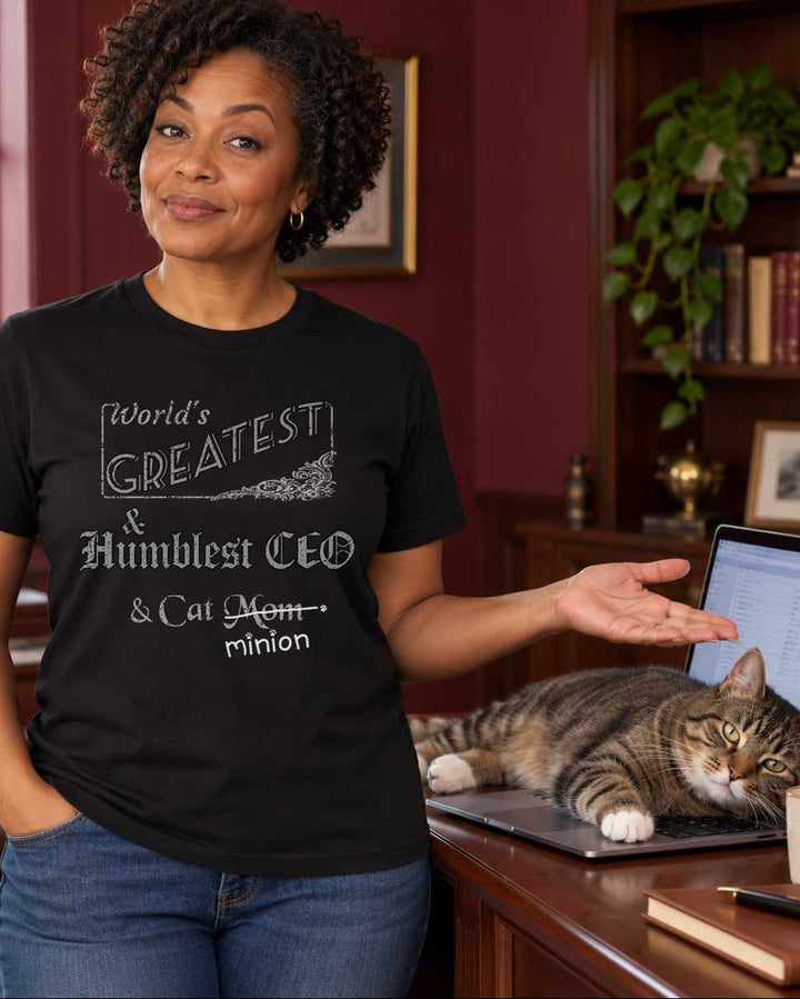Woman presenting a cat sprawled across a laptop in an office, wearing a black crew-neck t-shirt that reads, “World’s Greatest & Humblest CEO & Cat Mom” with “Mom” crossed out and “minion” handwritten below with paw prints.