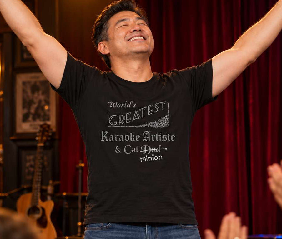 Man on a karaoke stage raising both arms as an audience applauds, wearing a black crew-neck t-shirt that reads, “World’s Greatest Karaoke Artiste & Cat Dad” with “Dad” crossed out and “minion” handwritten below with paw prints.