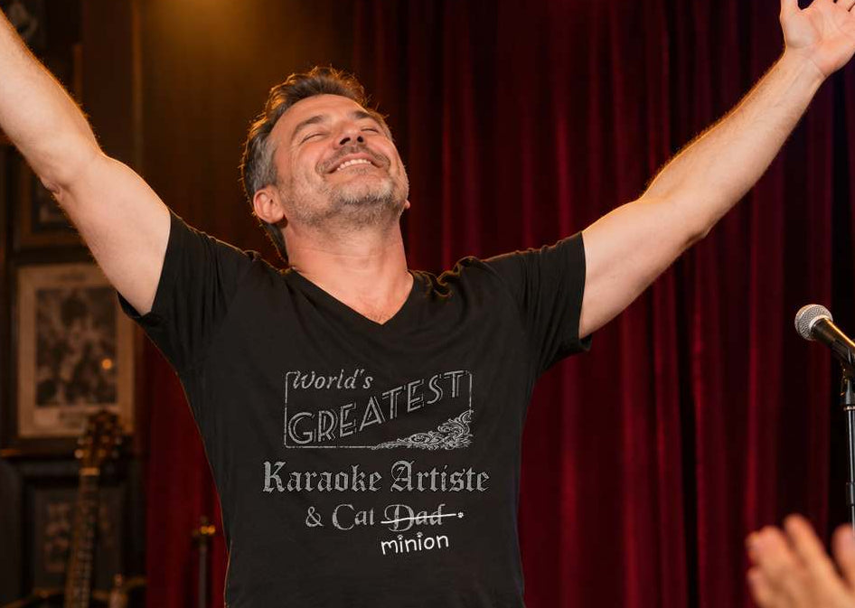 Man raising both arms in triumph onstage before an applauding crowd, wearing a black V-neck T-shirt reading “World’s Greatest Karaoke Artiste & Cat Dad.” “Dad” is crossed out, with “minion” handwritten beneath it and surrounded by cat paw prints.