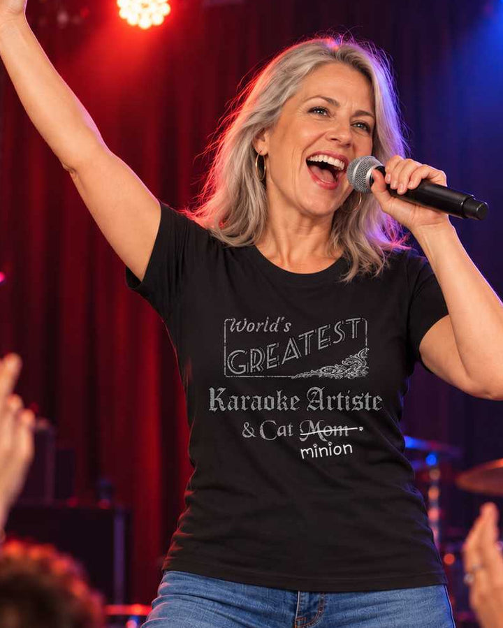 Woman singing into a microphone with one arm raised on a karaoke stage, wearing a black crew-neck t-shirt that reads, “World’s Greatest Karaoke Artiste & Cat Mom” with “Mom” crossed out and “minion” handwritten below with paw prints.