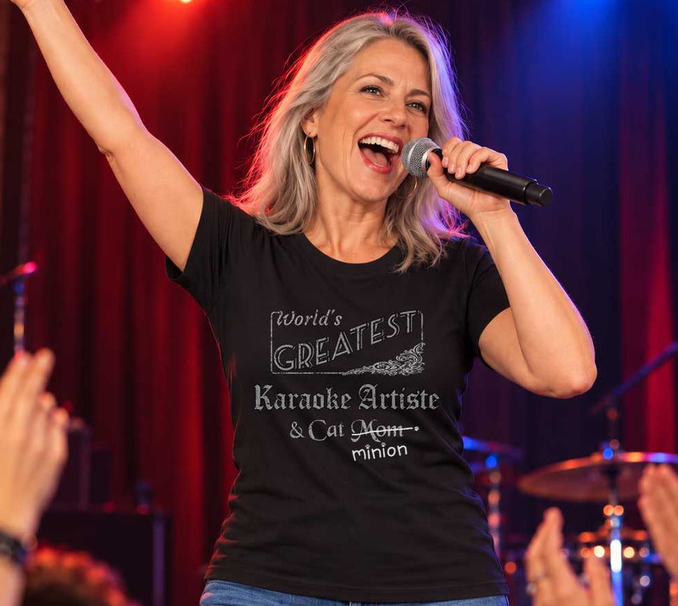 Woman singing into a microphone with one arm raised on a karaoke stage, wearing a black crew-neck t-shirt that reads, “World’s Greatest Karaoke Artiste & Cat Mom” with “Mom” crossed out and “minion” handwritten below with paw prints.