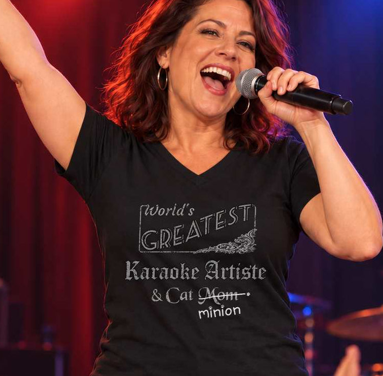 Woman singing into a handheld microphone with one arm raised, wearing a black V-neck T-shirt reading “World’s Greatest Karaoke Artiste & Cat Mom.” “Mom” is crossed out, with “minion” handwritten beneath it and surrounded by cat paw prints.