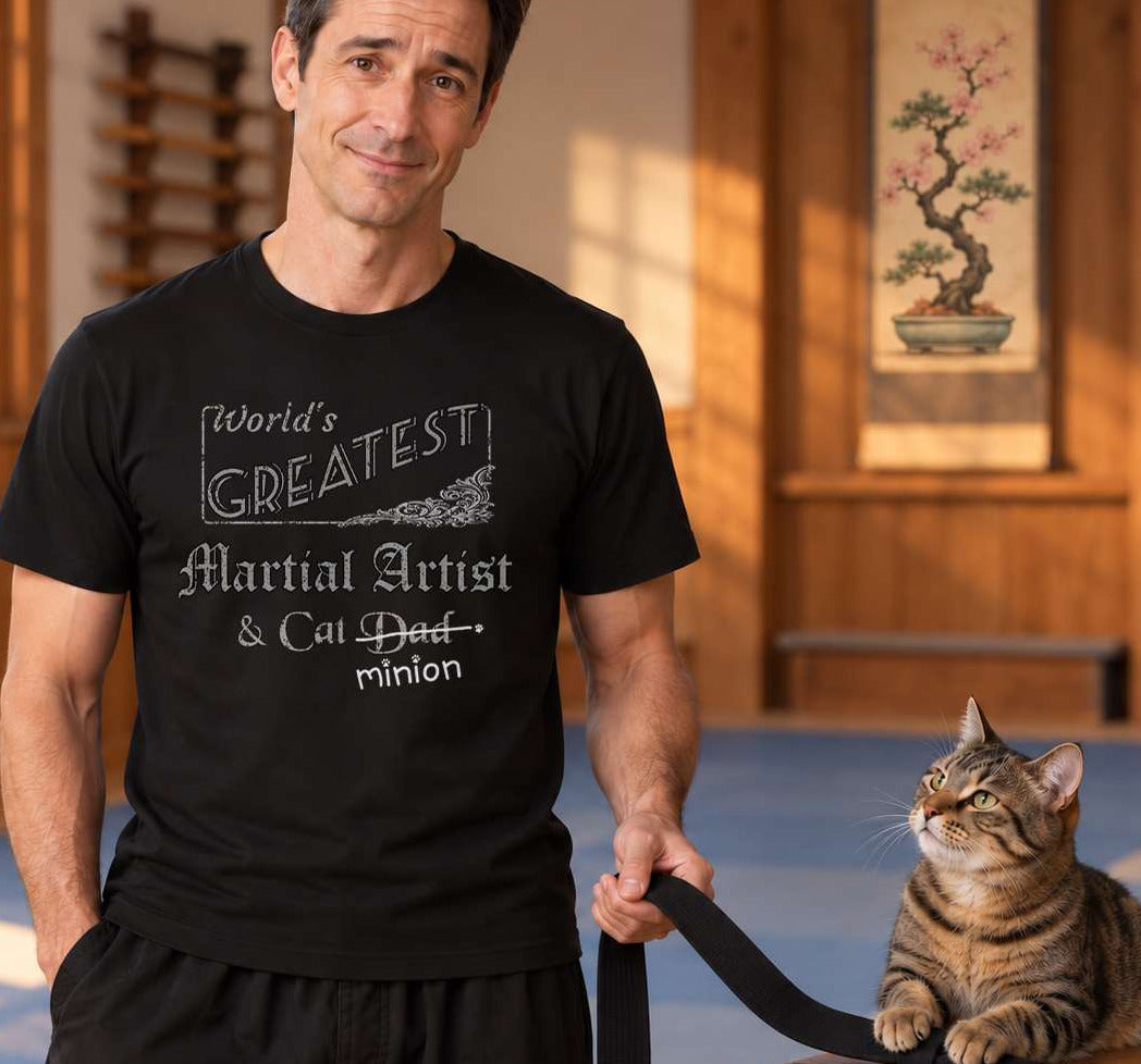 Man holding a martial-arts belt pinned by a cat on a dojo bench, wearing a black crew-neck t-shirt that reads, “World’s Greatest Martial Artist & Cat Dad” with “Dad” crossed out and “minion” handwritten below with paw prints.