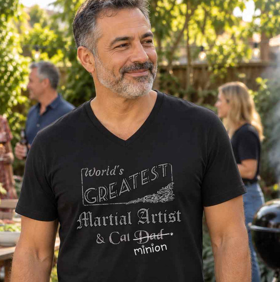 Man standing at an outdoor barbecue with friends, wearing a black V-neck T-shirt reading “World’s Greatest Martial Artist & Cat Dad.” “Dad” is crossed out, with “minion” handwritten beneath it and surrounded by cat paw prints.