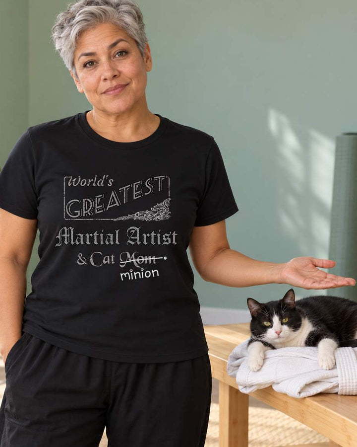 Woman presenting a cat sprawled on a folded training jacket in a home training room, wearing a black crew-neck t-shirt that reads, “World’s Greatest Martial Artist & Cat Mom” with “Mom” crossed out and “minion” handwritten below with paw prints.