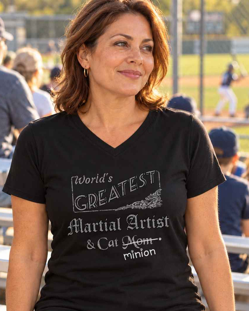 Woman sitting in the bleachers at a ballgame, wearing a black V-neck T-shirt reading “World’s Greatest Martial Artist & Cat Mom.” “Mom” is crossed out, with “minion” handwritten beneath it and surrounded by cat paw prints.
