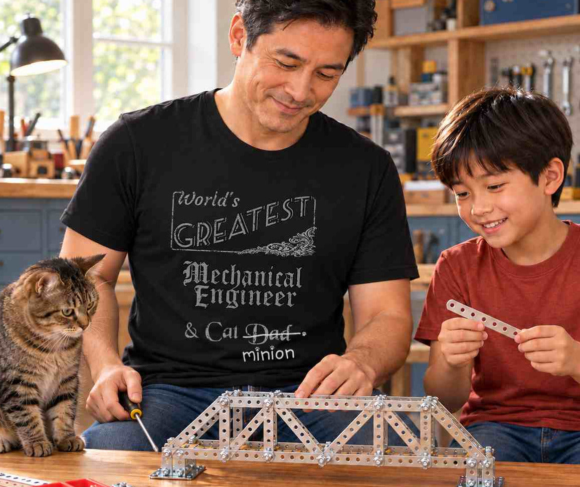 Man building a metal model bridge with a boy while a tabby cat supervises in a workshop, wearing a black crew-neck t-shirt that reads, “World’s Greatest Mechanical Engineer & Cat Dad” with “Dad” crossed out and “minion” handwritten below with paw prints.