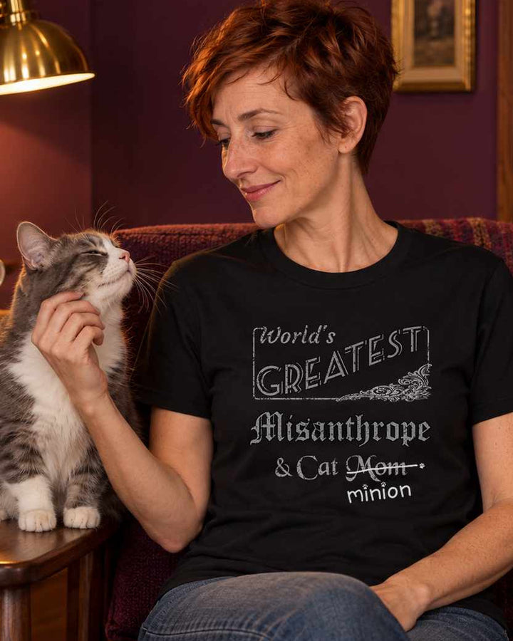 Woman scratching a grey-and-white cat’s cheek in a warmly lit plum reading nook, wearing a black crew-neck t-shirt that reads, “World’s Greatest Misanthrope & Cat Mom” with “Mom” crossed out and “minion” handwritten below with paw prints.